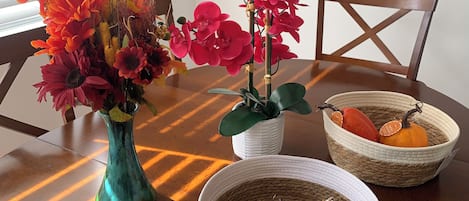 Une table à manger ornée d’un vase de fleurs, d’une plante en pot et de paniers tressés contenant des courges et une tomate.