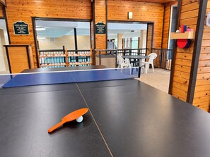 Mesa de pingue-pongue com raquete e bola, espaço de estar com cadeiras e parede de madeira com sinalização de área de comida e bebidas.