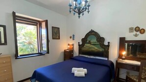 A bedroom with a large bed, a desk, a dresser, a chandelier, and a window with a view of greenery. - Relaxing Home Near the Best Beaches of Stintino (Stintino)