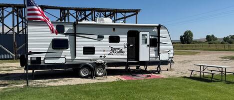 A Jayco Jayco 5X RV parked in a grassy area with a picnic table and a metal structure in the background.