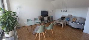 Een moderne woonkamer met een glazen eettafel, twee stoelen, een grijze bank, een salontafel en een televisie op een houten kast.