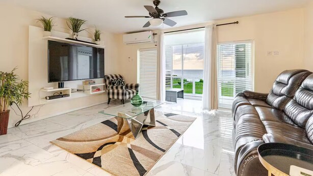 A modern living room with a flat-screen TV, a leather sofa, a glass coffee table, and a view of the outdoors through large windows.