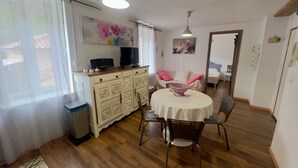 Un coin repas avec une table ronde, des chaises, un téléviseur posé sur un meuble et une fenêtre avec des rideaux.