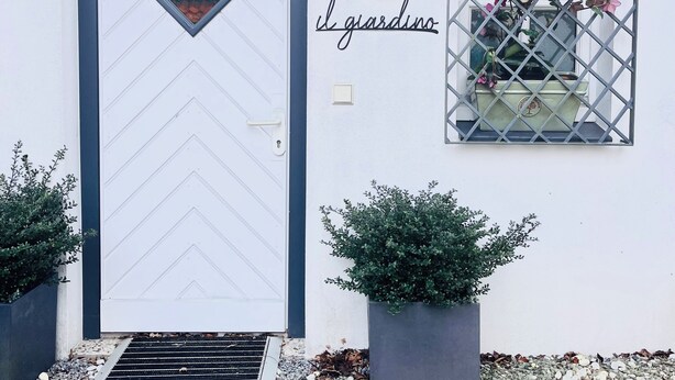 Un edifici blanc amb una porta apuntada d'estructura negra, una finestra petita amb reixeta, i el nom “il giardino” a la paret.