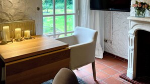 Habitació acollidora amb taula de fusta, una butaca blanca, una llar de foc, un televisor i una finestra gran amb vista a vegetació.