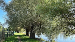 A grassy path beside a calm lake, with a wooden fence and a white plastic chair. - Vacation apartment Fischerhaus Havelberg (Havelberg)