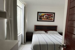 Relax in a cozy retreat with a large bed, bedside table, and a lamp, surrounded by a framed painting and natural light.