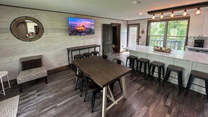 A modern kitchen with a large island, bar stools, a wooden dining table, and a TV mounted on the wall.