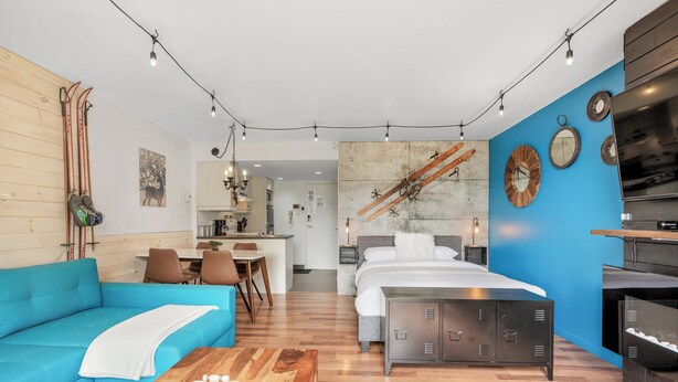 A modern hotel room with a blue accent wall, a wooden sofa, a bed with a grey headboard, a wooden coffee table, and a kitchen area visible in the background. - LOFT de la montagne- MSA (Saint-Ferréol-les-Neiges)