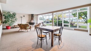 A spacious indoor area with a large table and chairs, a fireplace, and large windows offering a view of the outdoors. - LOFT de la montagne- MSA (Saint-Ferréol-les-Neiges)