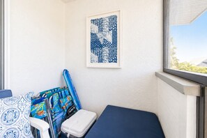 Ein kleiner Raum mit Fenster, einem gerahmten Kunstwerk, einer blauen Matratze, einem weißen Stuhl und einer bunten Tasche.