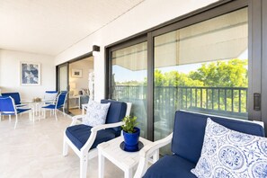 A balcony with white chairs and blue cushions, a glass door leading to a room, and a view of trees.