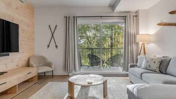 Ein modernes Wohnzimmer mit einem Flachbildfernseher, einem grauen Sofa, einem runden Couchtisch und einem großen Fenster mit Blick auf Bäume.
