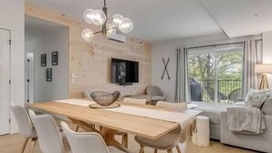 Ein modernes Wohnzimmer mit einem Holzesstisch, weißen Stühlen, einem Sofa und einem an der Wand befestigten Fernseher.