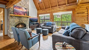A cozy log cabin interior with a stone fireplace, comfortable seating, and a large window overlooking greenery.