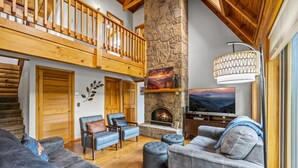 A spacious living room with a stone fireplace, wooden ceiling beams, and a large TV.
