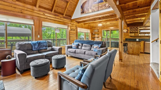 A spacious log cabin interior with a large living area, multiple sofas, ottomans, and a kitchen area.