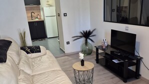 A modern living room with a white leather sofa, a round coffee table, a TV on a black stand, and a kitchen in the background. - L'Atelier T2 Cosy on the first floor (Saint-Gaudens)