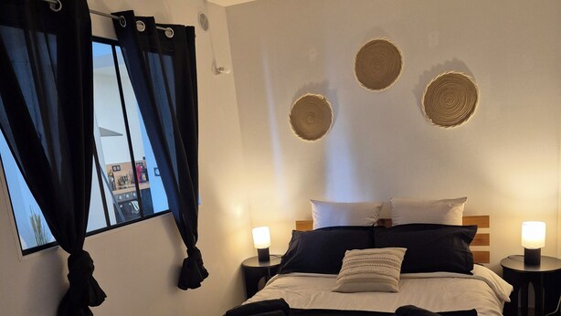 A bedroom with a wooden bed, black bedding, two bedside lamps, and a view of a kitchen through a window with black curtains. - L'Atelier T2 Cosy on the first floor (Saint-Gaudens)