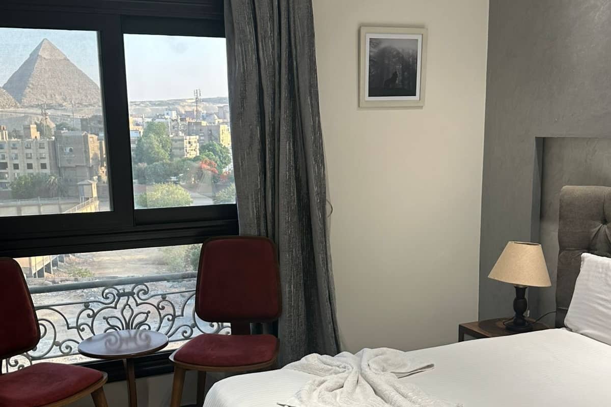 Habitación de hotel con una cama, dos sillas rojas, una mesita, una lámpara y vista a una pirámide.