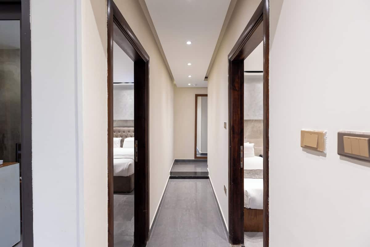 Un pasillo estrecho con una cama al final, una puerta a la izquierda y un interruptor de luz a la derecha.