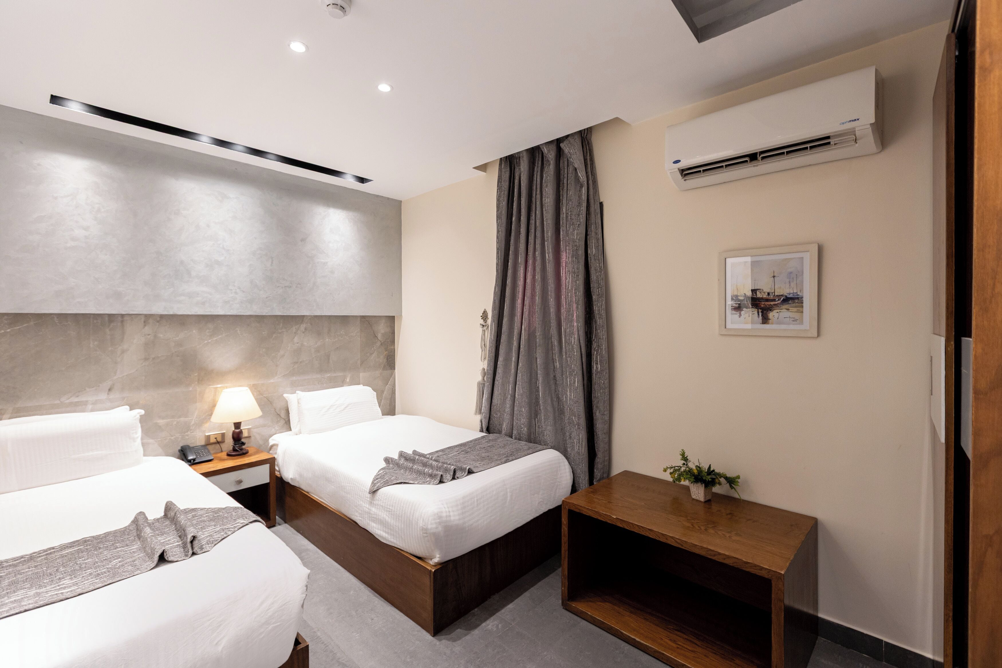 A modern hotel room with two beds, a bedside table with a lamp, a small wooden side table, and a potted plant.
