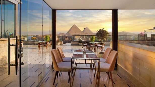 A rooftop terrace with a glass wall, a table with chairs, and a view of the pyramids. - pyramids Yard Hotel (Giza)