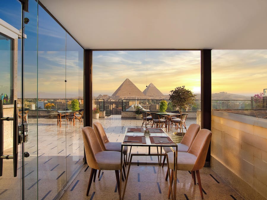 Terraza en la azotea con muro de cristal, una mesa con sillas y vistas a las pirámides.