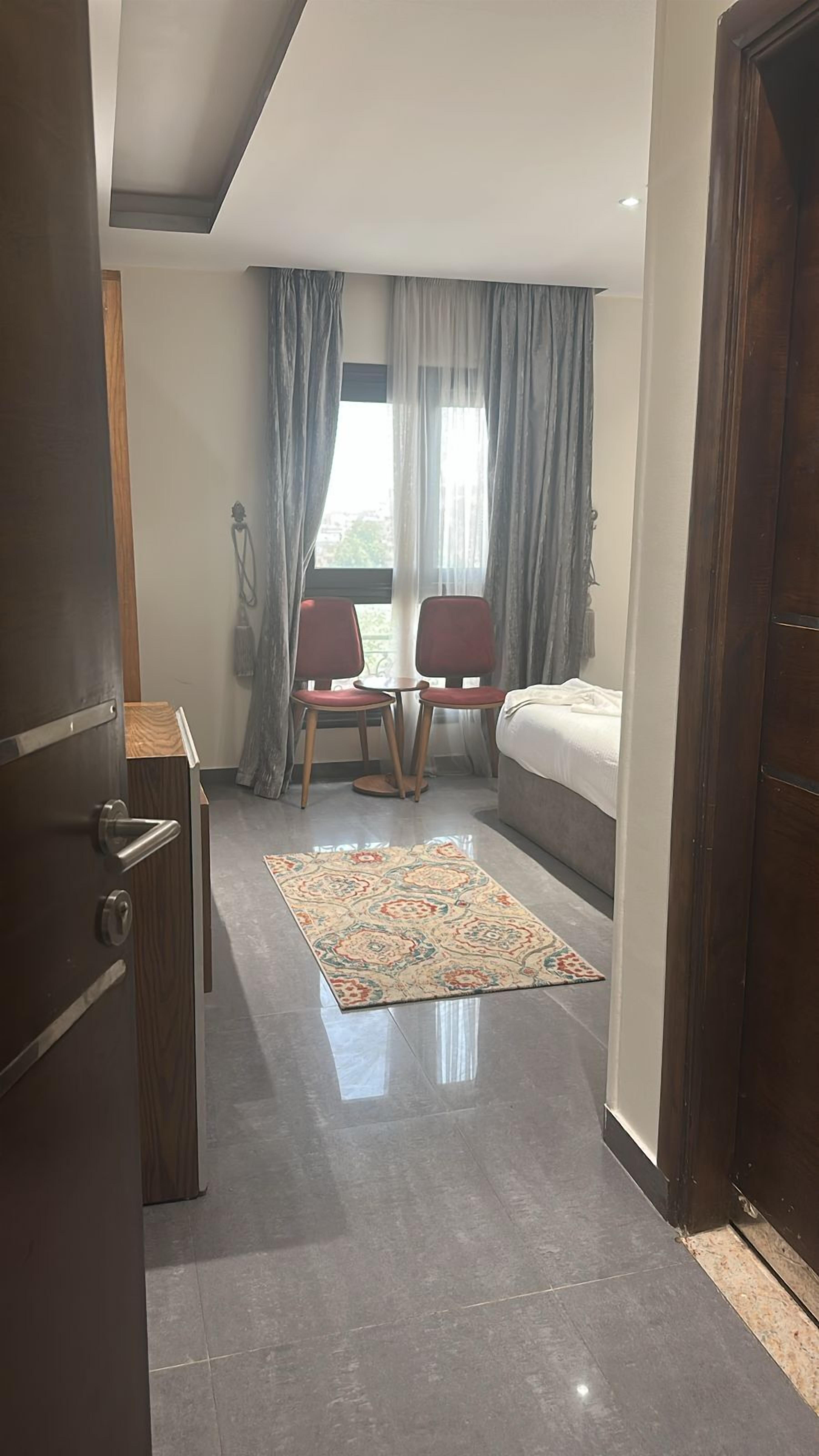 A hotel room with a bed, two chairs, a small table, a rug, and a window with curtains.