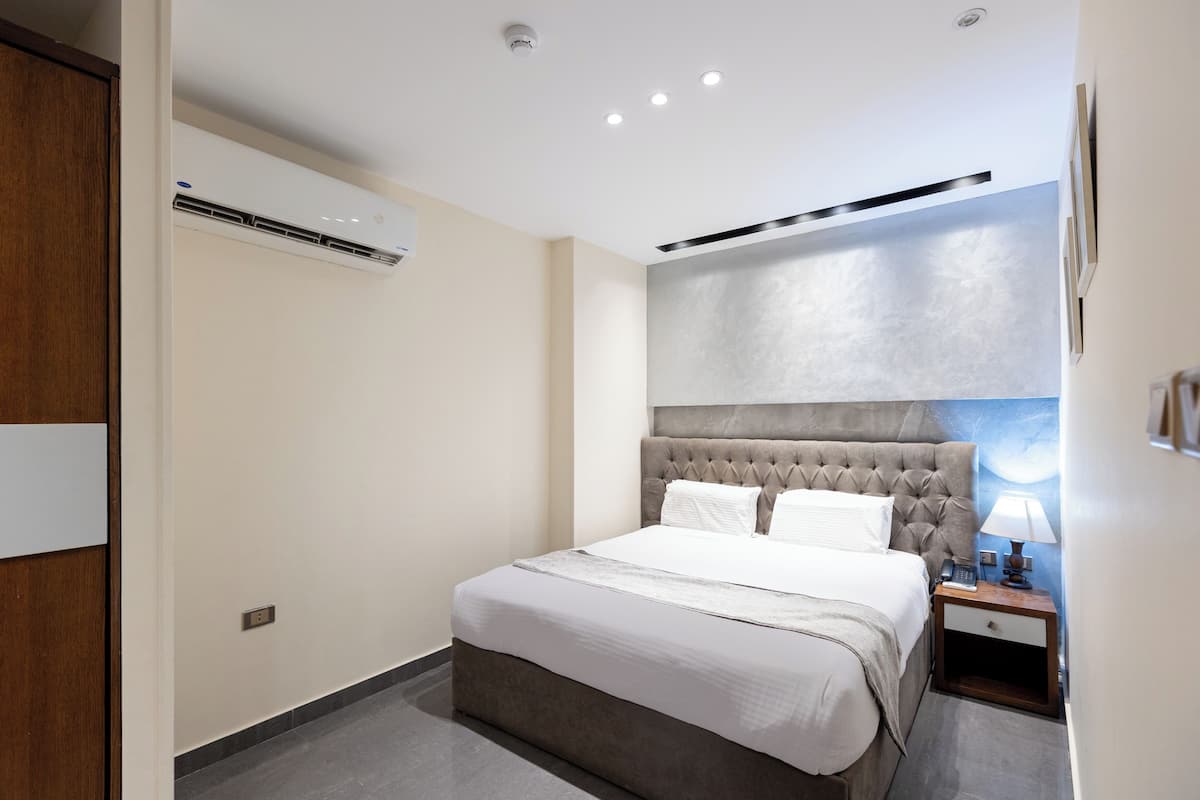 Una habitación de hotel moderna con una cama grande, una mesita de noche, aire acondicionado empotrado y luces de techo encastradas.