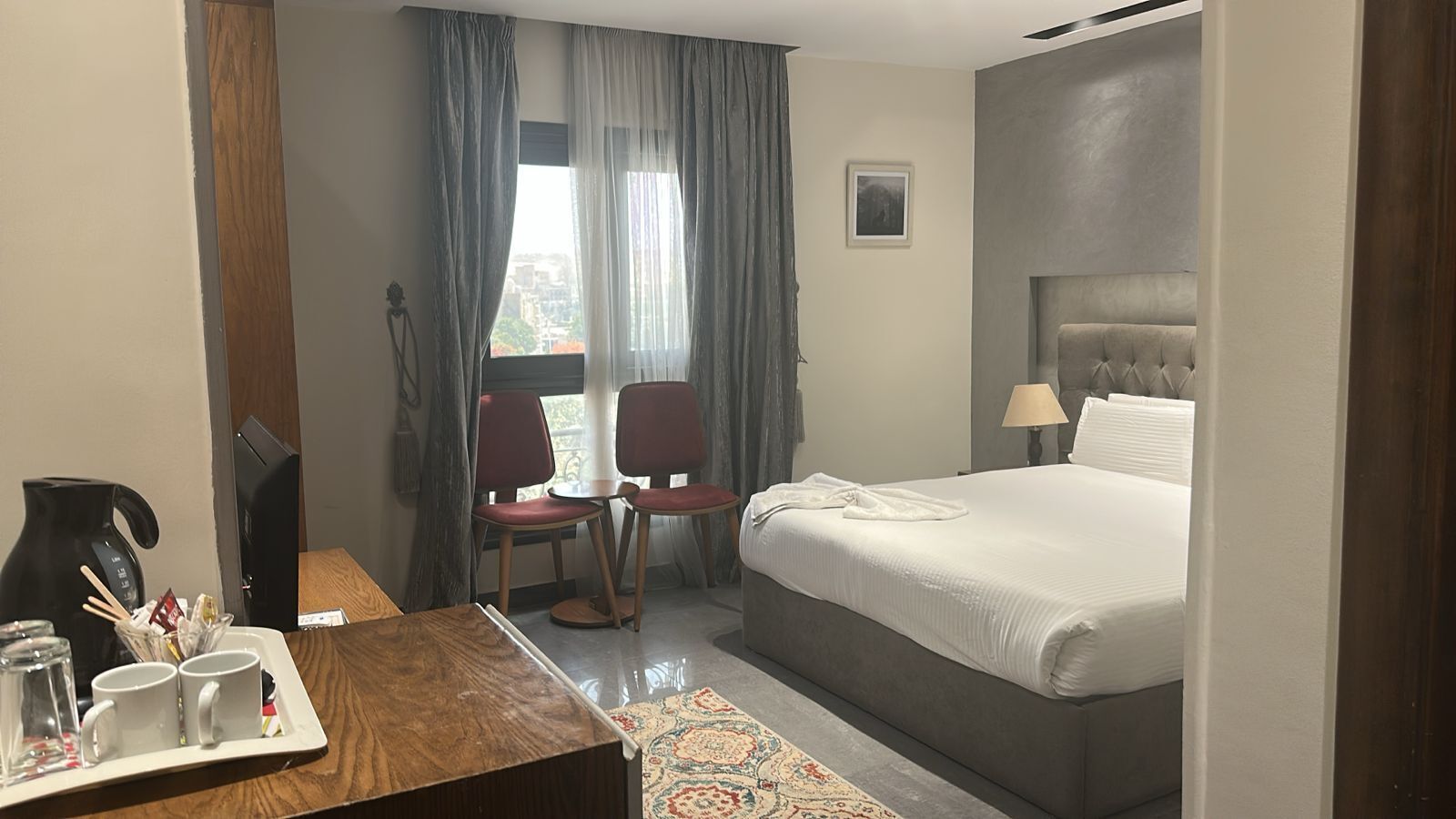 Hotellihuone, jossa on sänky, kaksi tuolia, työpöytä, jossa on kahvinkeitin, ja ikkuna, jossa on verhot.
