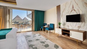 A modern hotel room with a large bed, a TV mounted on a marble wall, a balcony with a view of pyramids, and a seating area with chairs and a table. - pyramids Yard Hotel (Giza)
