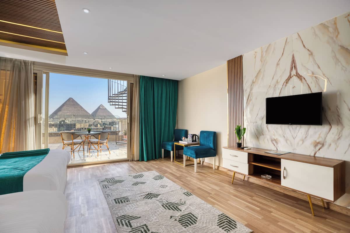 Una habitación de hotel moderna con una cama grande, un televisor montado en una pared de mármol, un balcón con vistas a las pirámides y una zona de estar con sillas y una mesa.