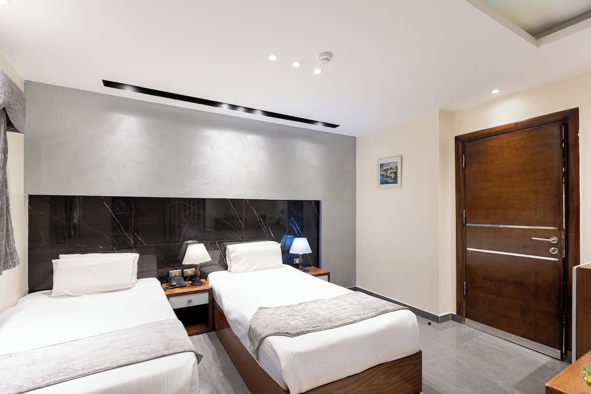 Habitación de hotel con dos camas, cabecera de mármol, lámparas de noche, puerta de madera y lámpara en el techo.
