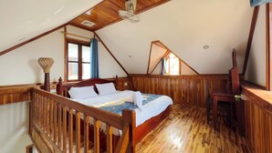 A bedroom with a sloped ceiling, wooden flooring, a bed with white bedding, a wooden chair, and a window with curtains. - BANSUAN Homes (Luang Prabang)