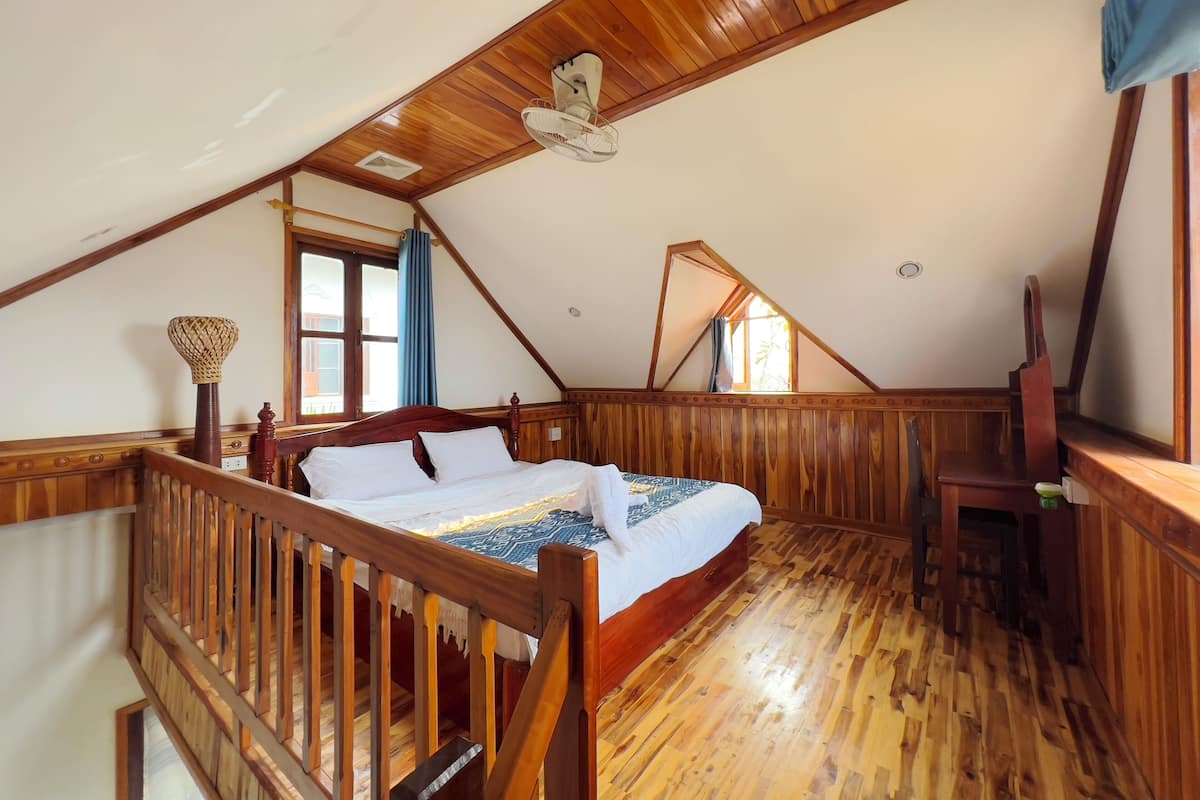 Une chambre avec un plafond en pente, un plancher en bois, un lit avec une literie blanche, une chaise en bois et une fenêtre avec des rideaux.