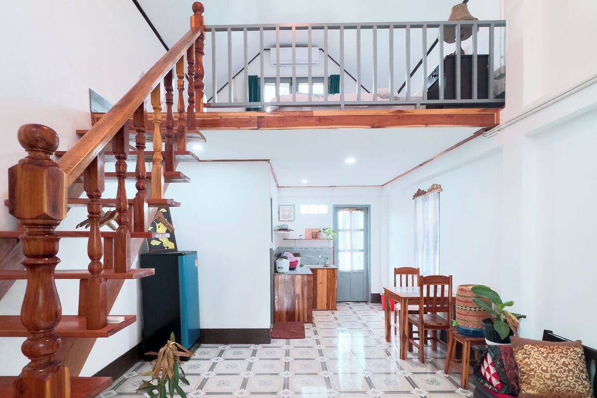 Un vaste espace de vie avec un escalier en bois menant à un niveau supérieur, un coin repas avec des chaises et une table, et une cuisine à l’arrière-plan.