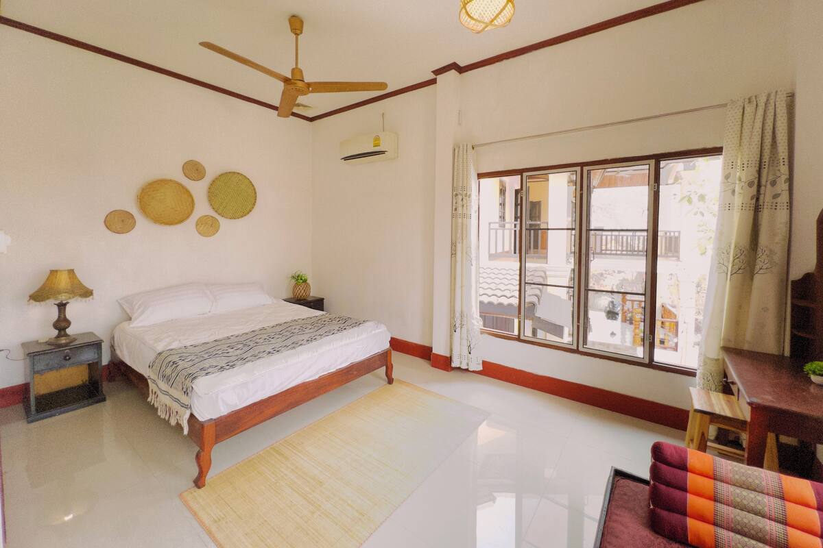 Une chambre à coucher comprenant un lit, une table de chevet, une lampe, un ventilateur de plafond, une fenêtre avec des rideaux et une vue sur l’extérieur.