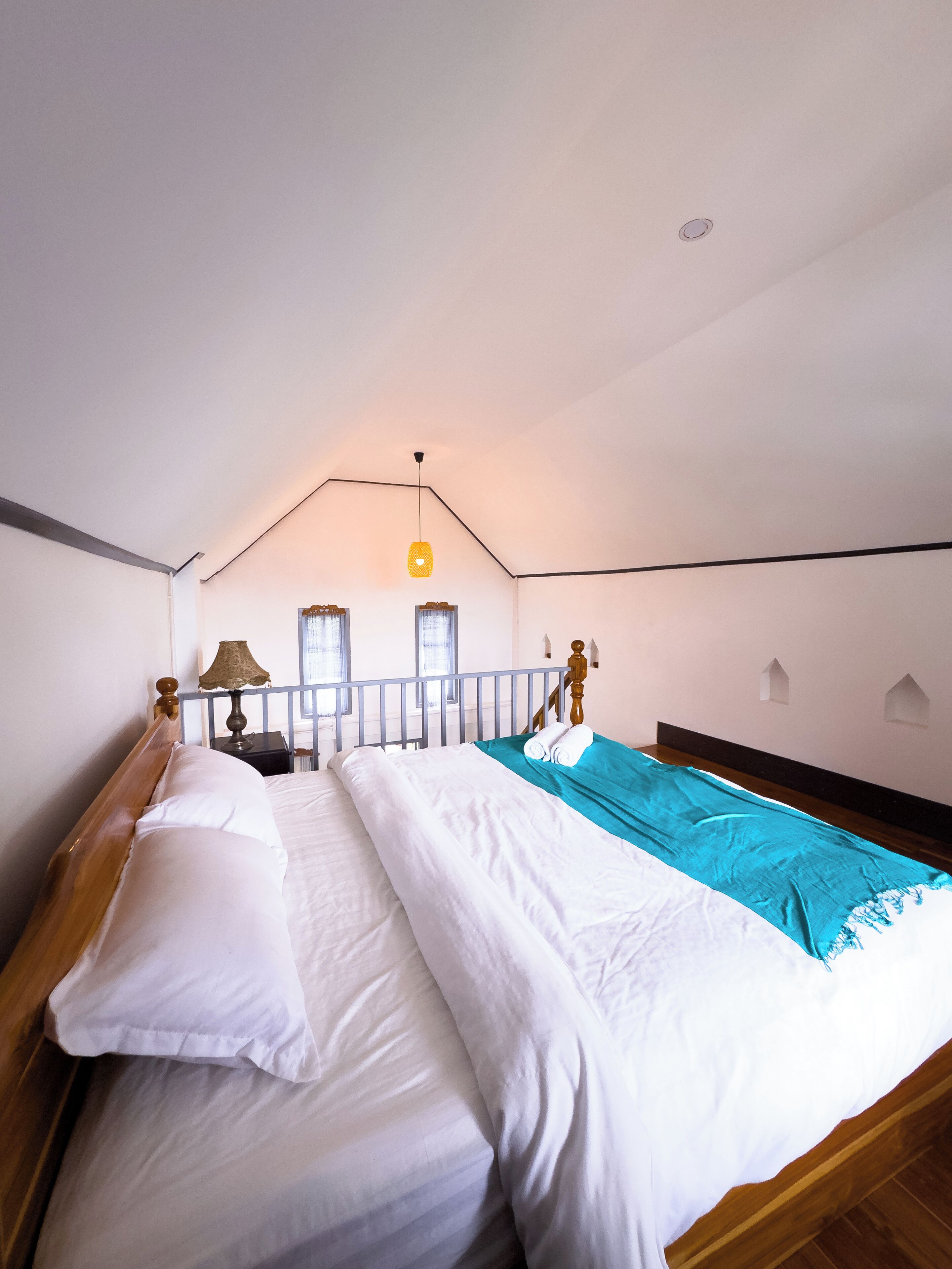 A bedroom with a wooden bed, white bedding, a teal blanket, a lamp, and a window with curtains.