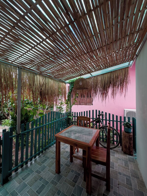 A covered patio area with a table and chairs, a green metal fence, and a pink wall. - BANSUAN Homes (Luang Prabang)