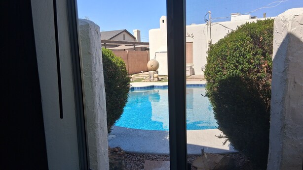 Vista de uma piscina pela janela, cercada por um muro branco e vegetação.