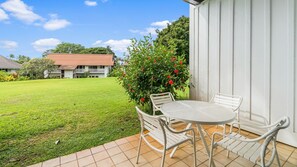 Eine Terrasse mit rundem Tisch und weißen Stühlen, ein kleiner Garten mit roten Blüten und ein weißes Gebäude im Hintergrund.