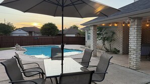 Ein Garten mit Schwimmbecken, einer überdachten Terrasse mit Tisch und Stühlen sowie einer Steinmauer.