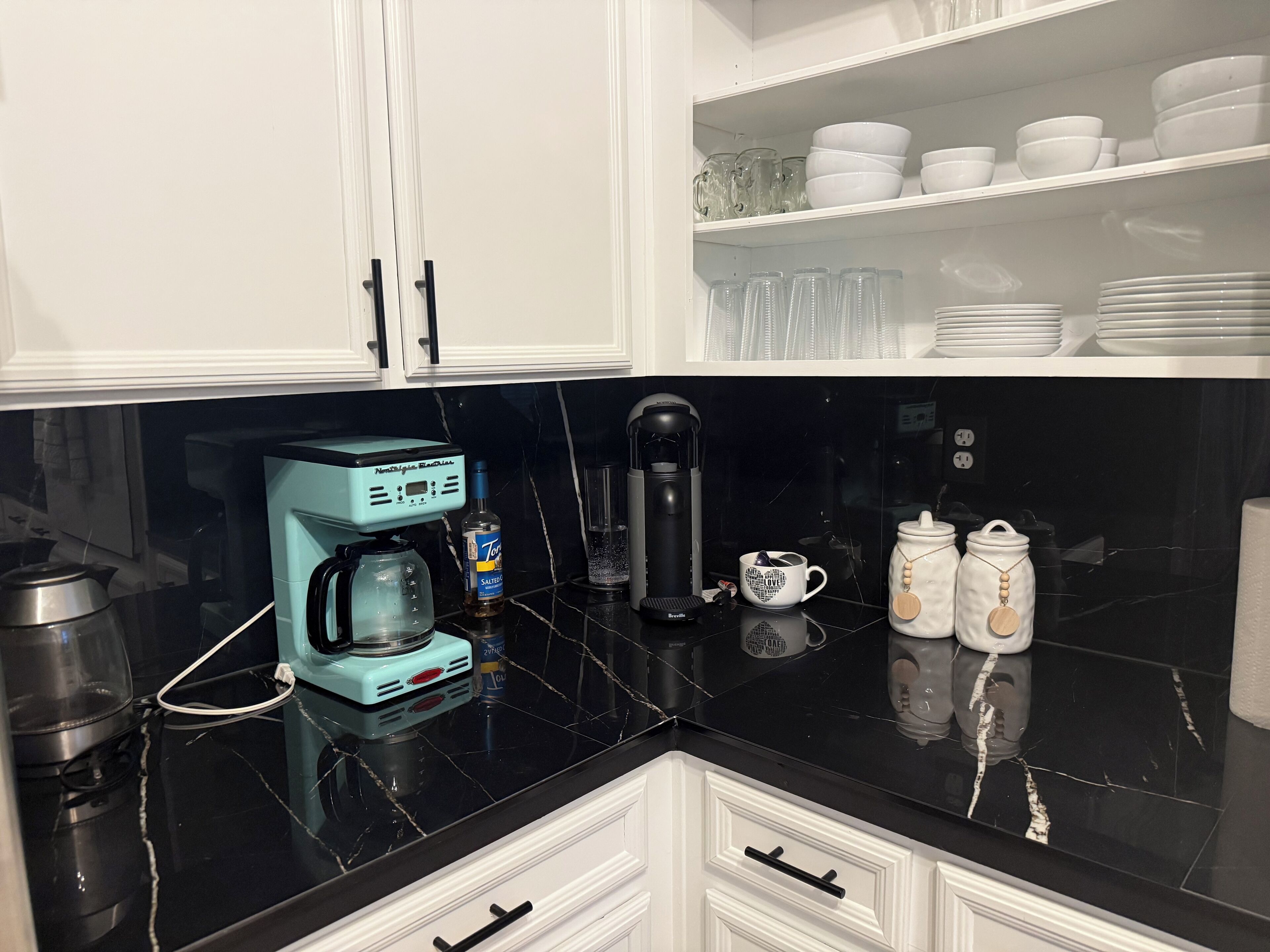 A kitchen with a black countertop, a coffee maker, a bottle of alcohol, and various kitchenware.