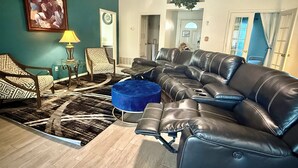 Ein Wohnzimmer mit Ecksofa, gemusterten Sesseln, einem blauen Hocker, einer Uhr und einem gerahmten Gemälde an der Wand.
