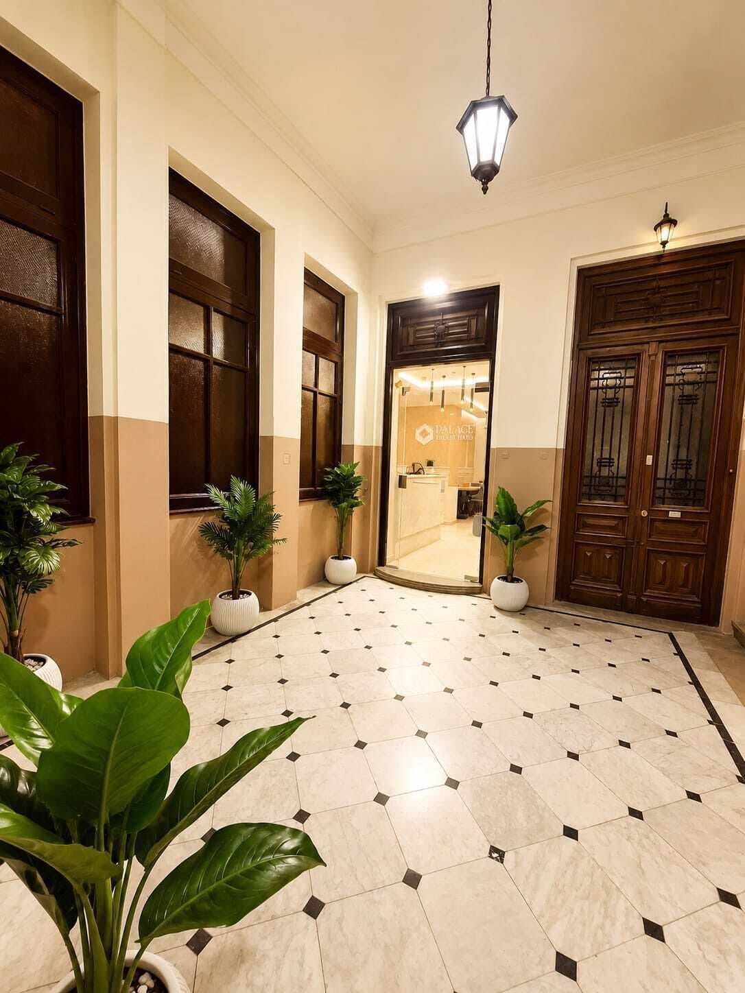 Un hall de hotel con suelo de baldosas, plantas en macetas, una puerta de madera y una lámpara colgante.
