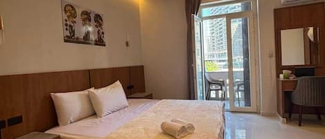 A hotel room with a large bed, a desk, and a chair. There is a window with curtains, a door, and an air conditioning unit.