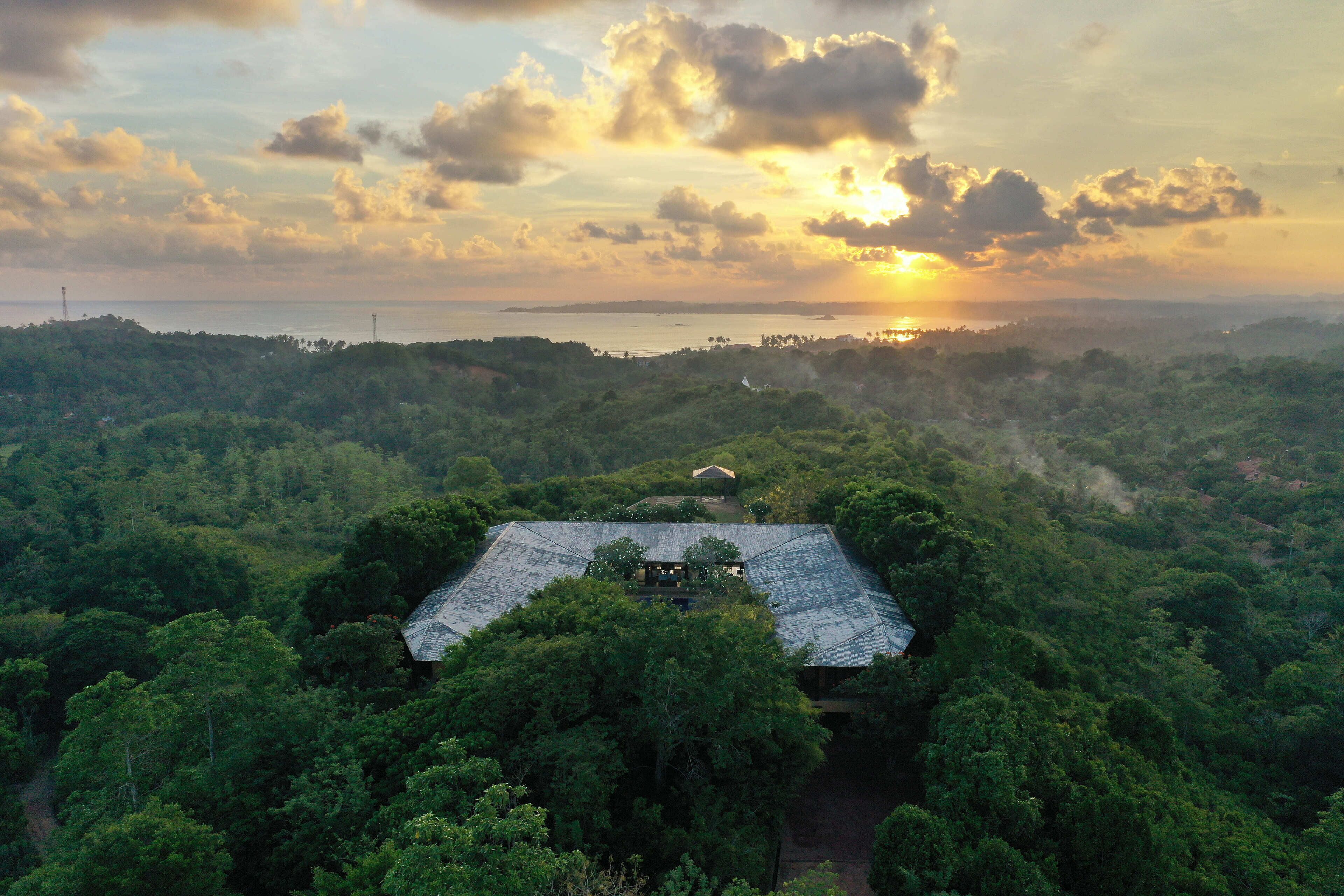 A house surrounded by dense forest with a clear view of the sunset over the ocean.