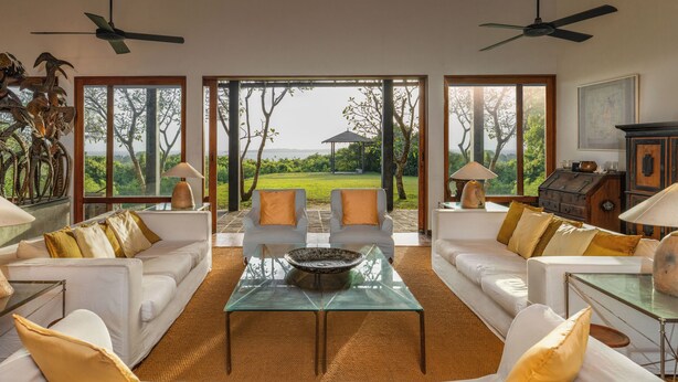 A spacious living area with a glass coffee table, white sofas, and large windows offering a view of a lush garden.