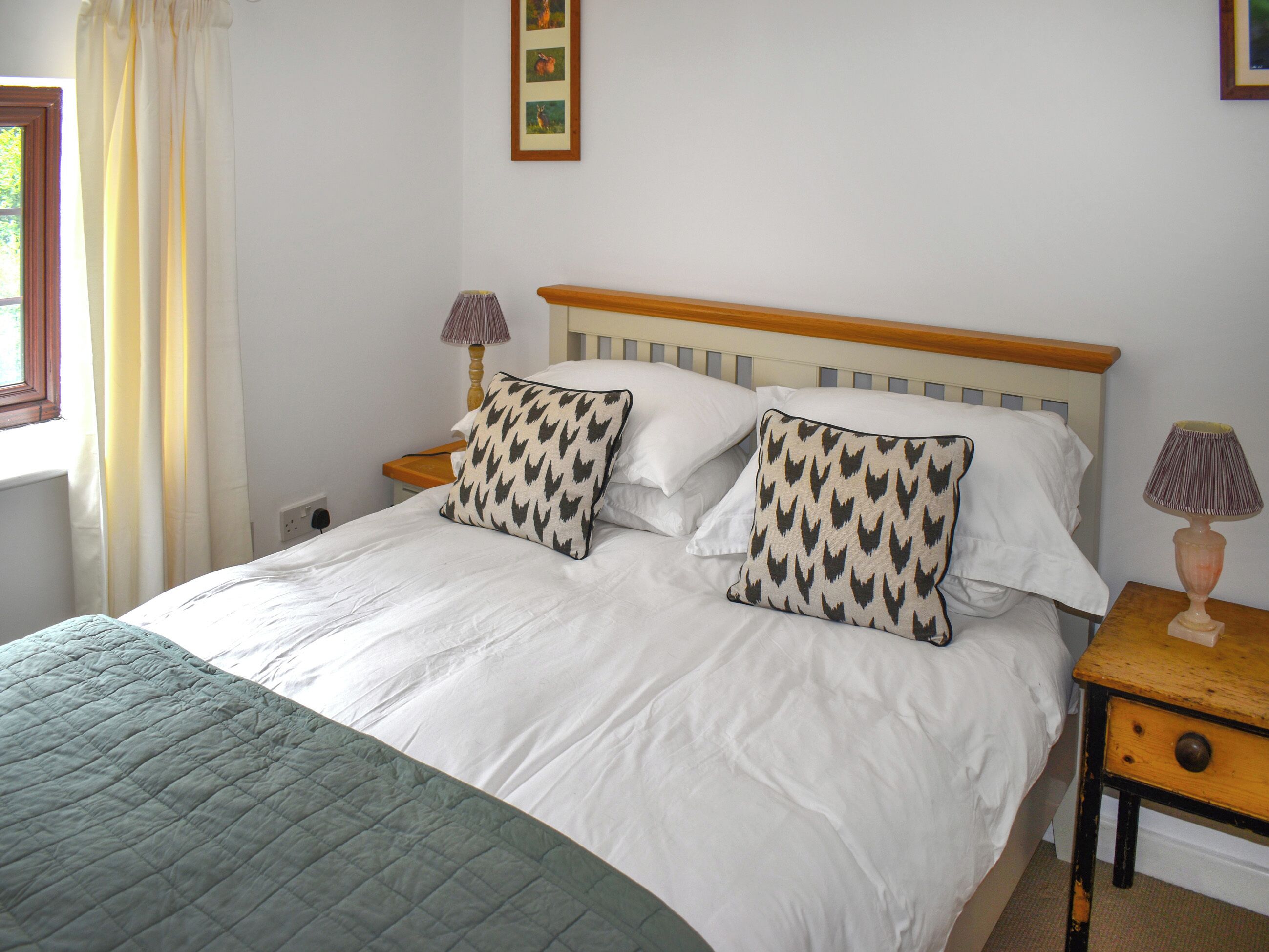Una cama bien tendida con almohadillas estampadas, una lámpara de noche y un cuadro enmarcado en la pared.